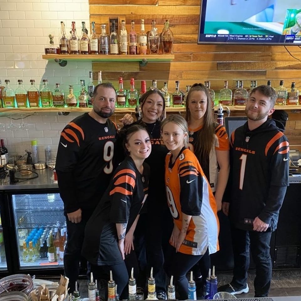 Five restaurant staff members pose behind a bar stocked with liquor bottles, wearing Cincinnati Bengals jerseys.