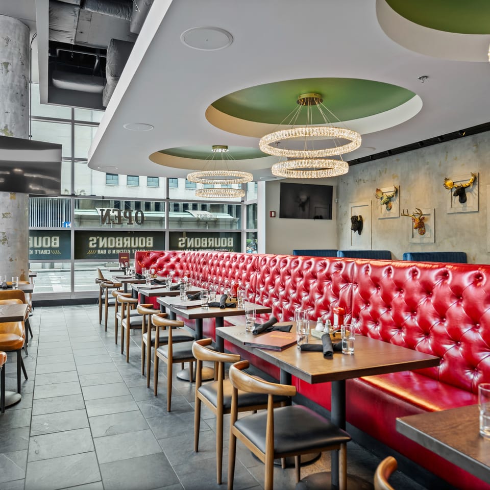 Modern restaurant dining room with red tufted booths, crystal ring chandeliers, set tables, and decorative animal-head wall sculptures.