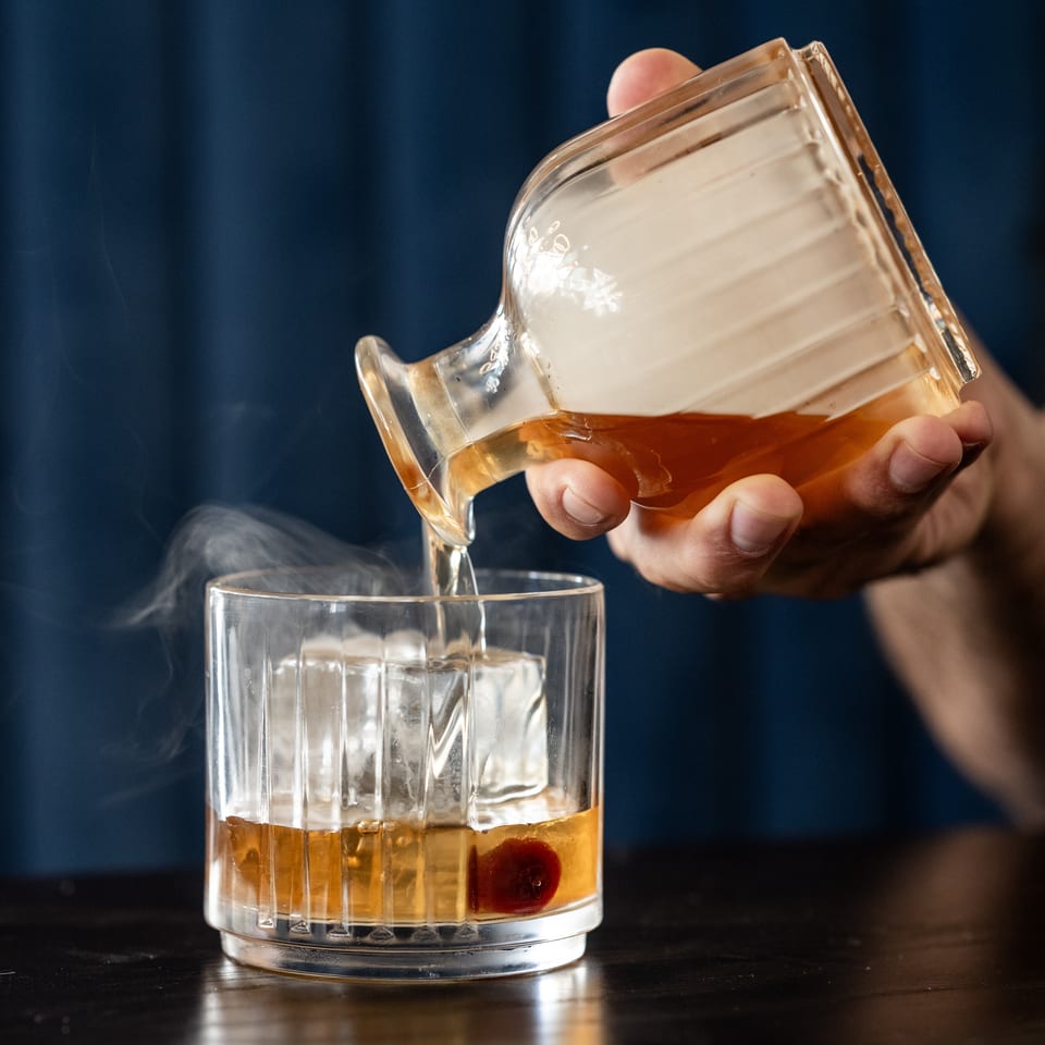 Bartender pours amber liquor from a glass pitcher into an ice-filled tumbler garnished with a cherry.