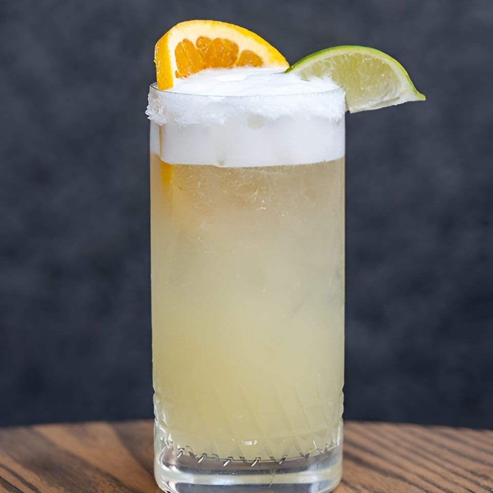 Pale citrus cocktail in a tall glass, topped with frothy foam and garnished with orange and lime wedges on a wooden table.