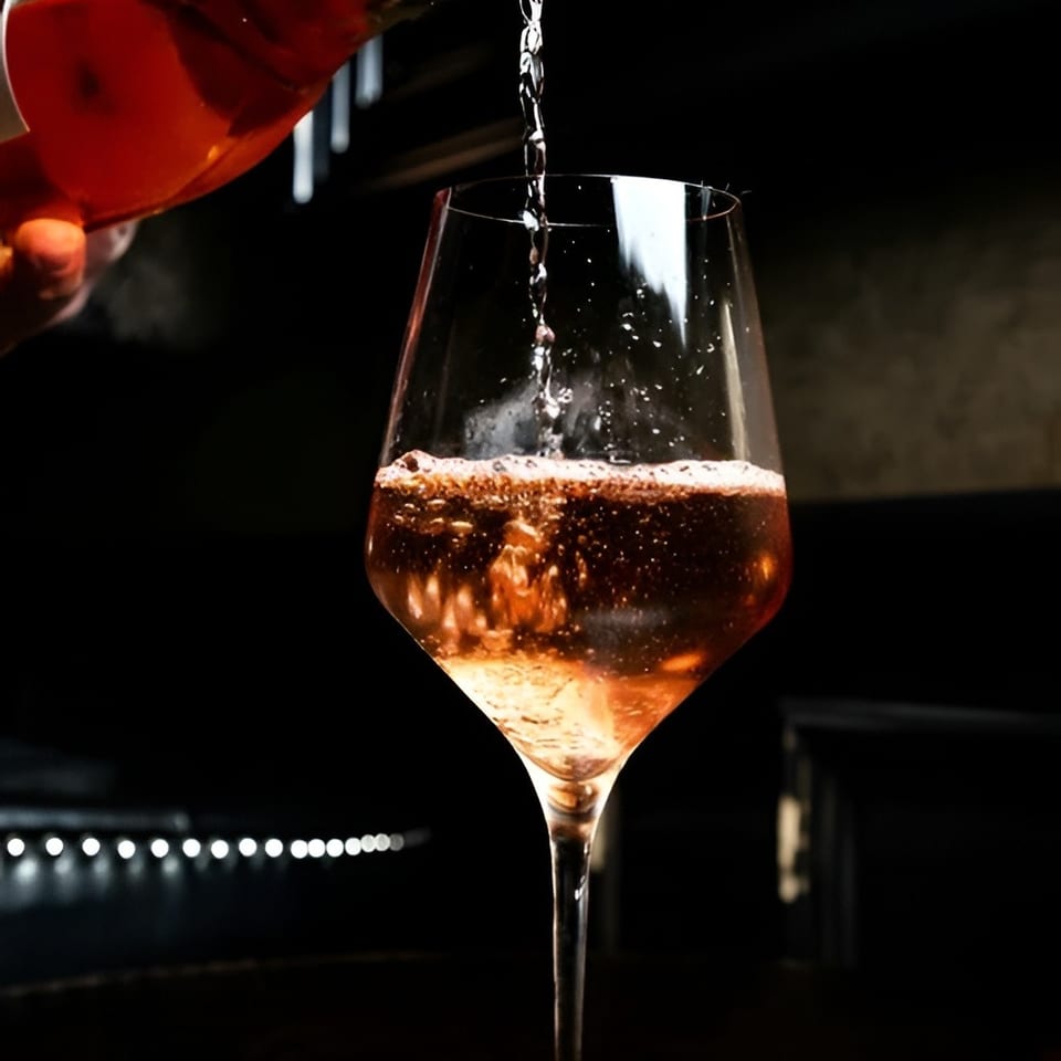 Red sparkling beverage pours from a bottle into a stemmed wine glass against a dark, warmly lit background.