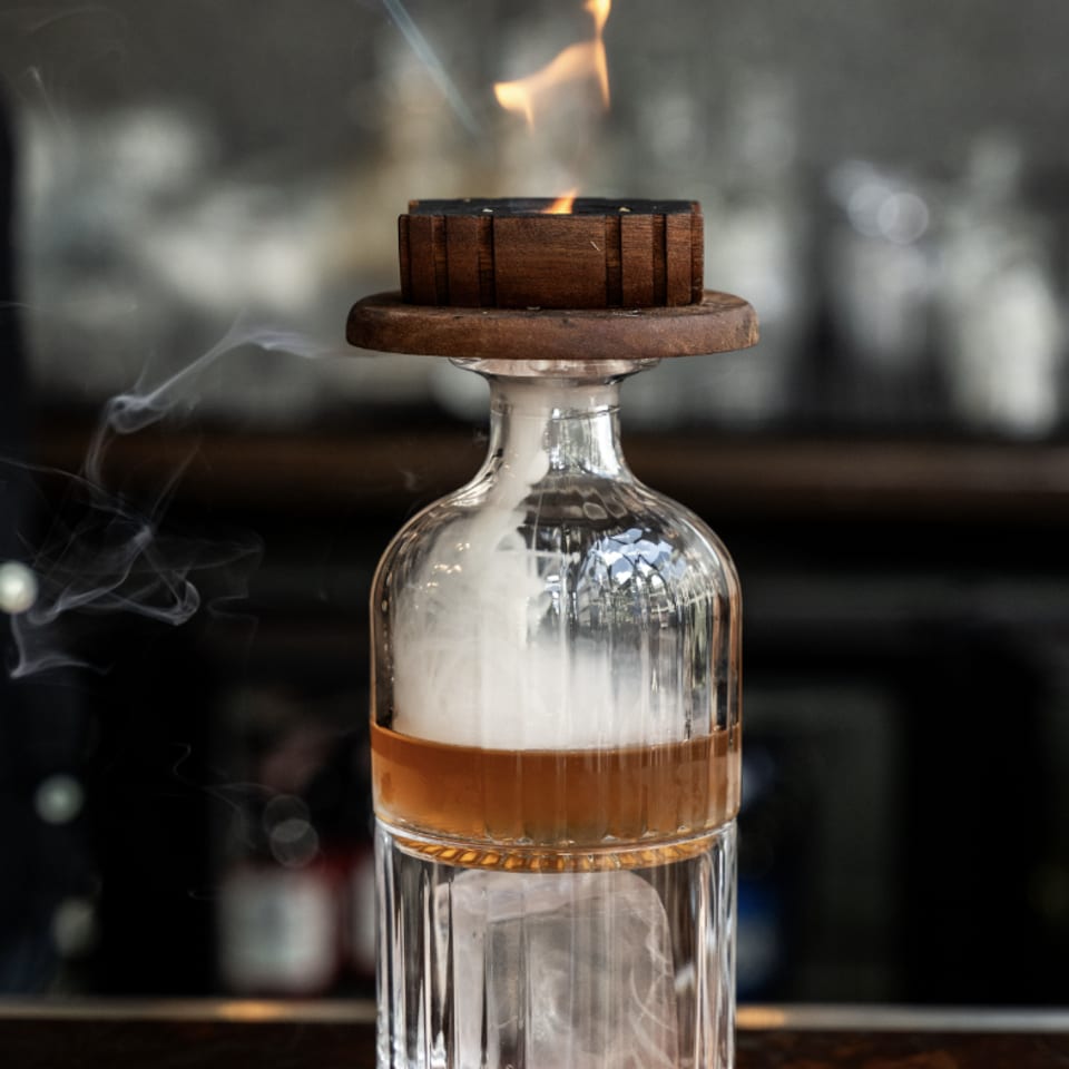 A bartender uses a torch to smoke a cocktail in a unique glass with a wooden top.