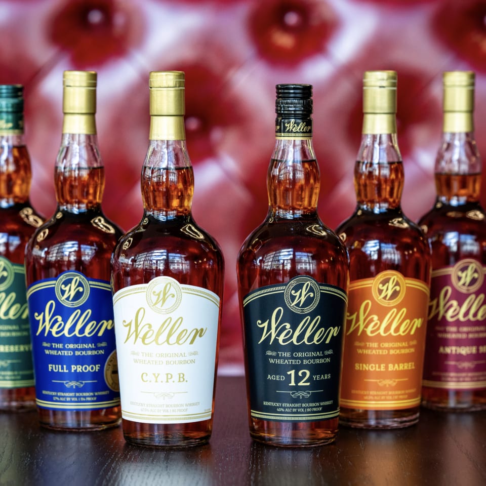 Six bottles of Weller bourbon whiskey, each with different colored labels, on a dark wooden surface.
