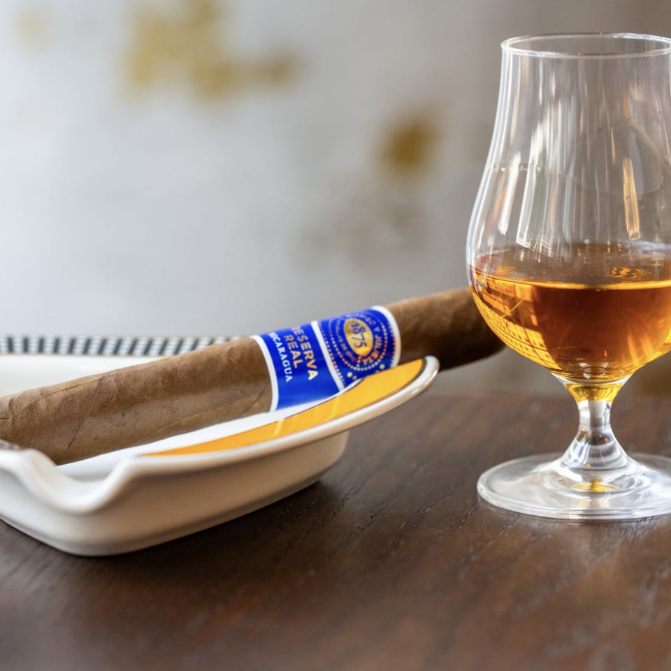 A cigar with a blue band rests in a white ashtray next to a glass of amber liquor.