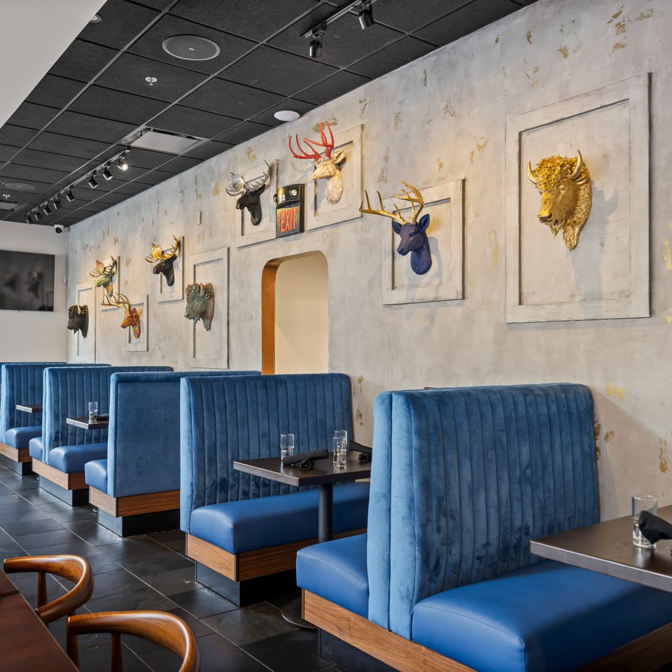 Restaurant dining room with blue velvet booths, wood tables, and colorful decorative animal heads mounted on a textured wall.