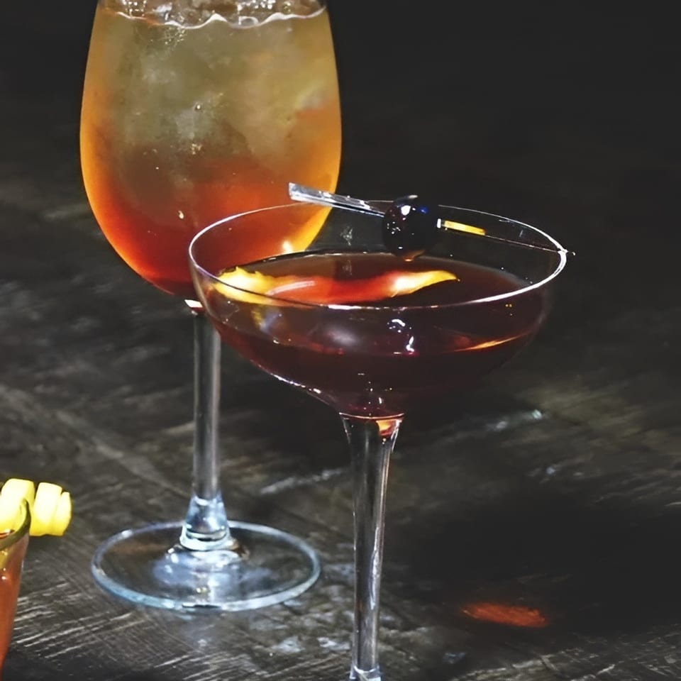 Two cocktails sit on a dark wooden table: an amber drink with ice and a deep-red coupe garnished with citrus.