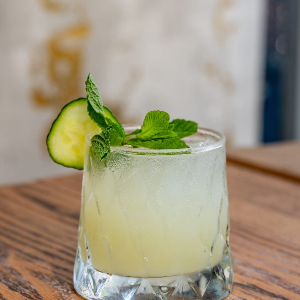 Pale green cucumber-mint drink in a chilled glass, garnished with cucumber slices and fresh mint on a wooden table.