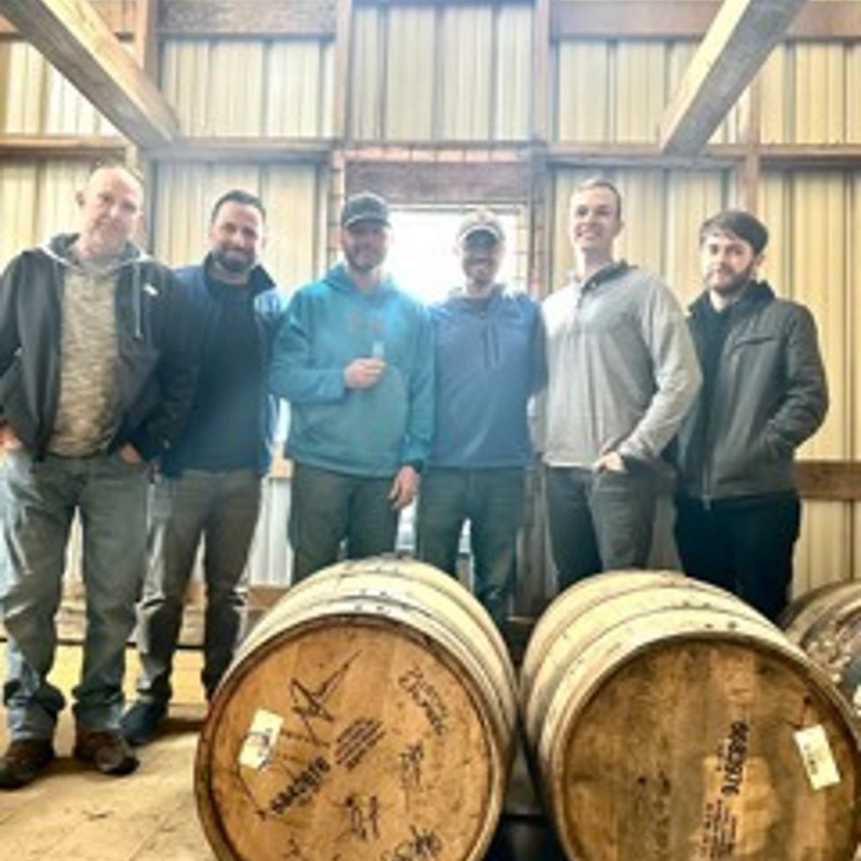Six men pose behind two wooden barrels inside a rustic barn with exposed beams and corrugated metal walls.