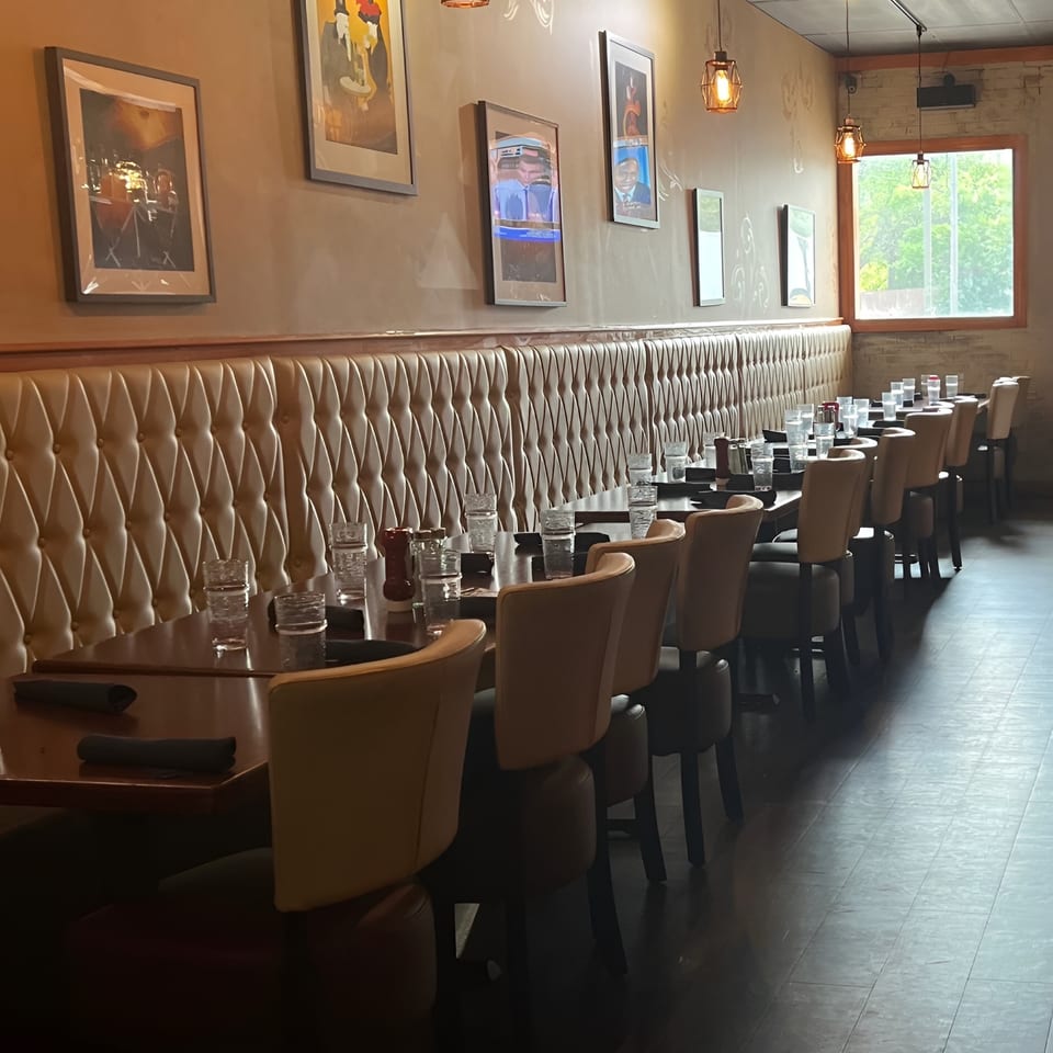 Warmly lit restaurant dining room with tufted banquette seating, set tables, framed artwork, pendant lights, and large windows.