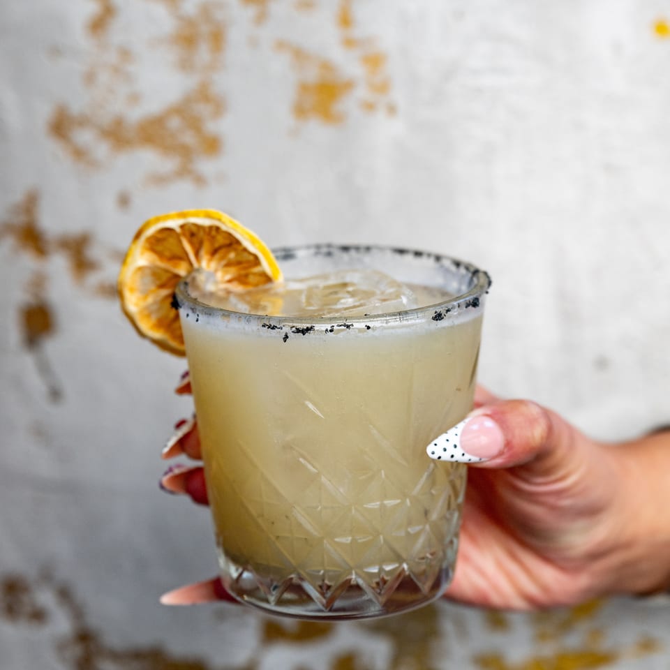 A pale citrus cocktail in a cut-glass tumbler with a black-salt rim and dried orange garnish, held by a person.