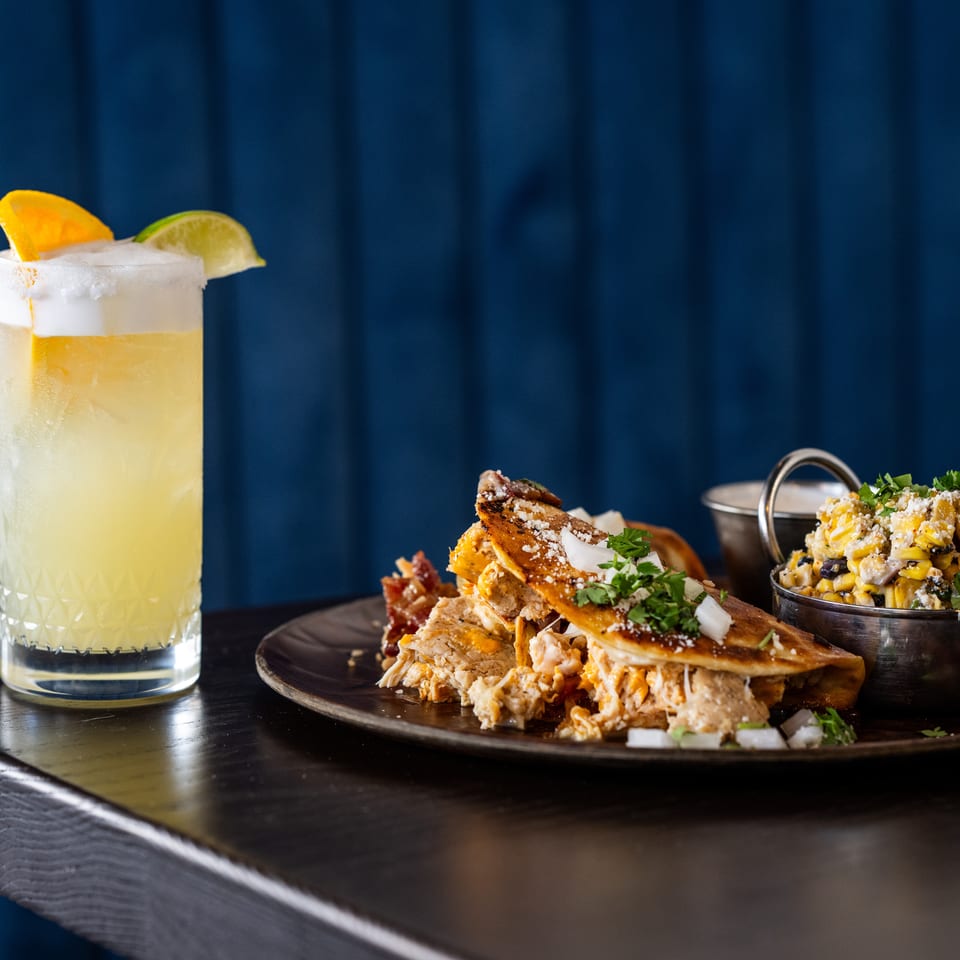 Frosty citrus cocktail with orange and lime garnishes beside loaded quesadillas and a bowl of corn salad on a dark table.