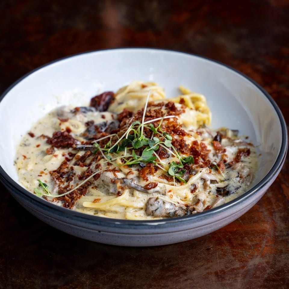 Creamy pasta with mushrooms, sun-dried tomatoes, crispy bacon bits, and microgreens in a white bowl.