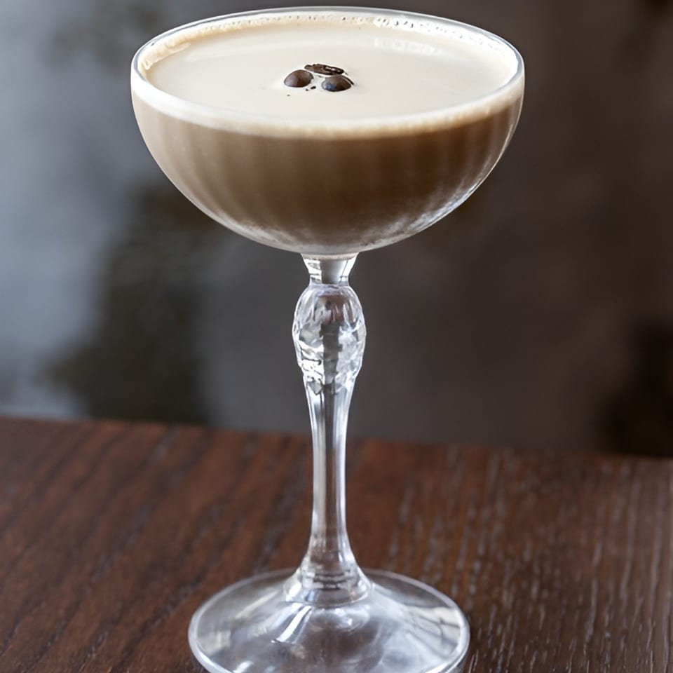 Creamy coffee cocktail topped with coffee beans, served in a stemmed coupe glass on a wooden table.