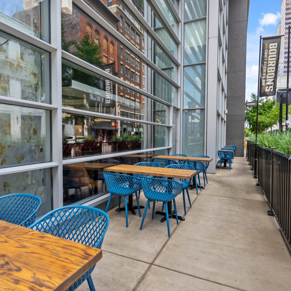 Bourbon’s Craft Kitchen and Bar outdoor patio with wooden tables, blue chairs, greenery, and an urban city backdrop.