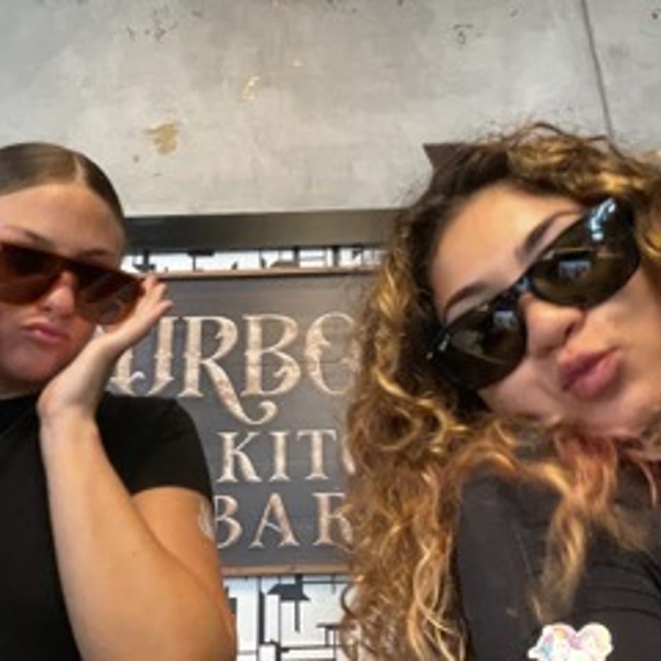 Two women wearing sunglasses pose indoors in front of an Urban Kitchen Bar sign.