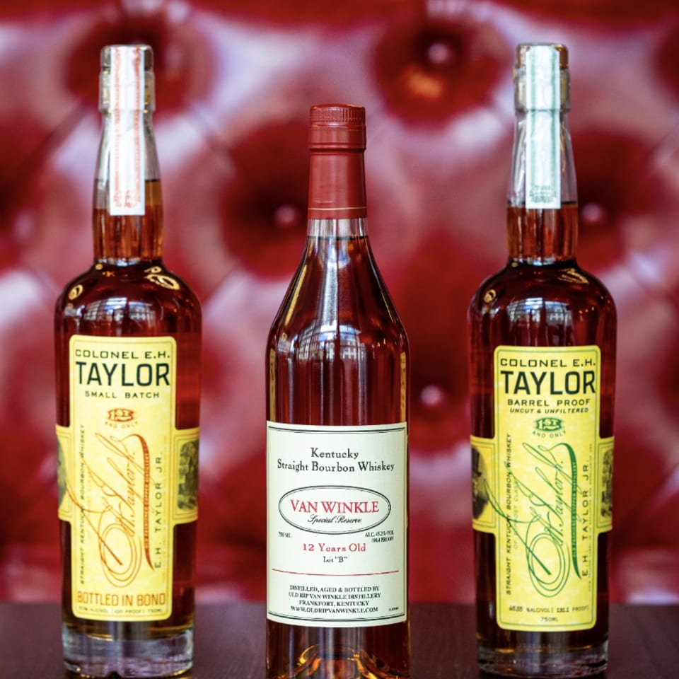 Three bottles of bourbon, including Colonel E.H. Taylor and Van Winkle, on a dark table.